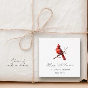 May include: A white card with a red cardinal bird perched on a branch. The card reads "Mary Williams In Loving Memory 1947-2025".