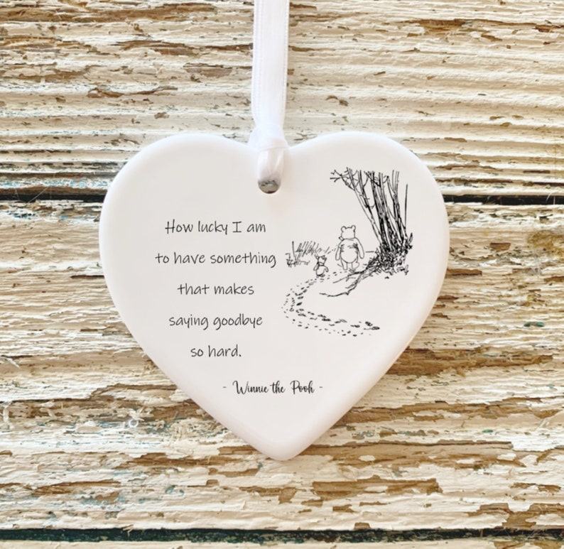 May include: A white heart-shaped ornament with a ribbon for hanging. The ornament features a Winnie the Pooh illustration and quote: "How lucky I am to have something that makes saying goodbye so hard." The quote is attributed to Winnie the Pooh.