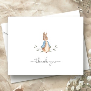 May include: A white thank you card with a watercolor illustration of a brown rabbit wearing a blue jacket and surrounded by green leaves. The text "thank you" is written in black cursive.