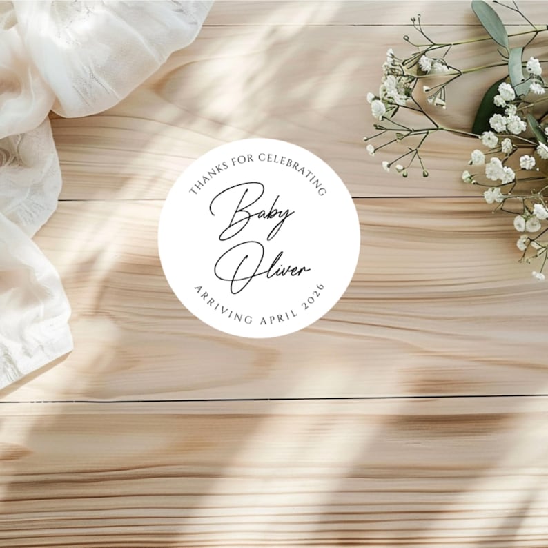 Puede incluir: Una pegatina circular blanca con el texto "THANKS FOR CELEBRATING Baby Oliver ARRIVING APRIL 2026" en escritura negra. La pegatina est&aacute; sobre una superficie de madera clara, con tela blanca y peque&ntilde;as flores blancas en el fondo.