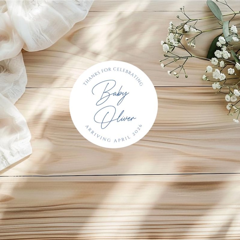 Puede incluir: Una pegatina circular blanca con texto azul que dice "Gracias por celebrar Baby Oliver Llegando Abril 2026". La pegatina est&aacute; sobre una superficie de madera marr&oacute;n claro, con flores blancas y tela en el fondo.