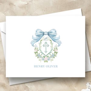 May include: A white card with a blue watercolor bow and floral wreath surrounding a silver cross. The text "HENRY OLIVER" is printed below the wreath.