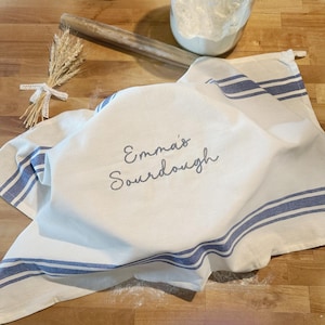 May include: A white kitchen towel with blue stripes and the embroidered text Emma's Sourdough. The towel is draped over a bowl, accompanied by a wooden rolling pin and a small bundle of dried wheat stalks tied with a white lace ribbon.