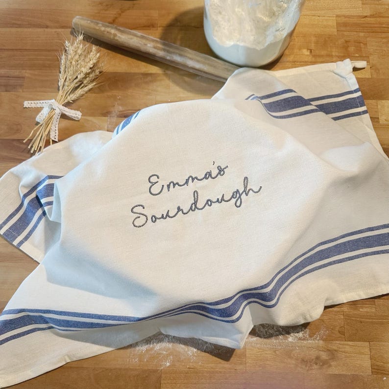 Puede incluir: Un pa&ntilde;o de cocina blanco con rayas azules y el texto bordado Emma's Sourdough. El pa&ntilde;o est&aacute; colocado sobre un cuenco, acompa&ntilde;ado de un rodillo de madera y un peque&ntilde;o manojo de tallos de trigo secos atados con una cinta de encaje blanca.