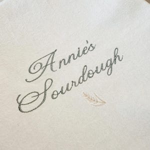May include: A cream-colored fabric surface featuring dark gray embroidered cursive text that reads Annie's Sourdough. Below the text is a small, light tan embroidered wheat stalk design. The fabric has a subtle, textured, woven appearance.