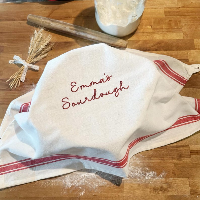 Puede incluir: Un pa&ntilde;o de cocina blanco con rayas rojas y el texto bordado en rojo Emma's Sourdough. El pa&ntilde;o cubre un cuenco, acompa&ntilde;ado de un rodillo de madera, un tarro de cristal lleno de harina y un peque&ntilde;o manojo de espigas de trigo secas.