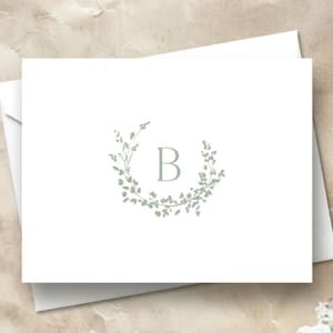 May include: A white card with a green leafy wreath design and the letter "B" in the center.