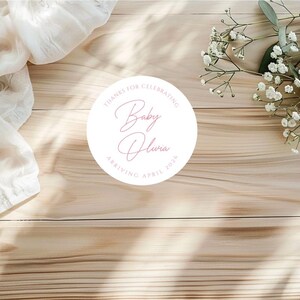 Puede incluir: Una pegatina circular blanca con texto rosa que dice "Gracias por celebrar Baby Olivia Llegada abril 2026". La pegatina est&aacute; sobre una superficie de madera clara, con tela blanca y flores blancas en el fondo.