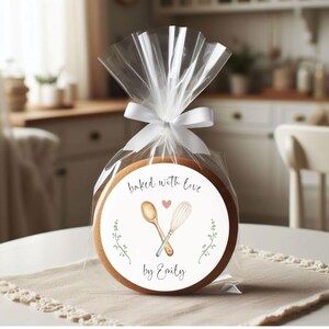 Puede incluir: Una galleta redonda marr&oacute;n con una pegatina blanca que dice "baked with love by Emily". La pegatina tiene un coraz&oacute;n rosa y un dibujo de una cuchara de madera y un batidor.