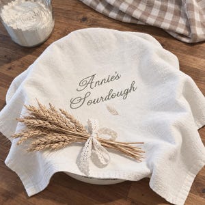 May include: An off-white kitchen towel featuring the embroidered text Annie's Sourdough in a cursive font, accented by a small wheat stalk icon. A bundle of dried wheat stalks tied with a white lace ribbon rests on top of the fabric.