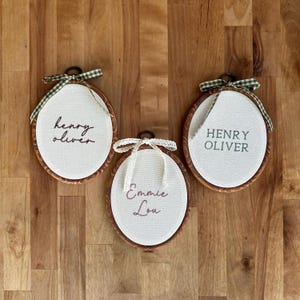 May include: Three oval embroidery hoops with wooden frames containing white fabric. The left hoop features the name Henry Oliver in dark script, the center hoop has Emmie Lou in pink script with a lace bow, and the right has Henry Oliver in green block letters with a green gingham bow.