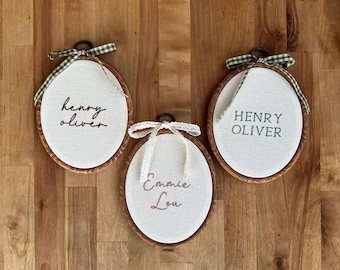Puede incluir: Tres bastidores de bordado ovalados con marcos de madera y tela blanca. El bastidor izquierdo muestra el nombre Henry Oliver en letra cursiva oscura, el central Emmie Lou en rosa con un lazo de encaje, y el derecho Henry Oliver en letras verdes con un lazo de cuadros vichy verde.