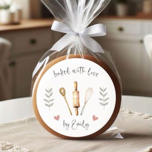 Puede incluir: Una pegatina redonda blanca con el texto "baked with love by Emily" y una ilustraci&oacute;n de acuarela de un rodillo, un batidor y una cuchara de madera. Dos corazones rosas est&aacute;n a cada lado del texto.