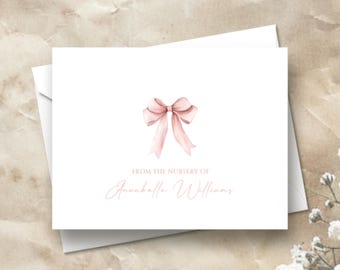 May include: White stationery card with a watercolor illustration of a pink bow. The text "FROM THE NURSERY OF" and a name are printed in a delicate script font. The card is placed on a white envelope.