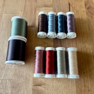 May include: A collection of ten sewing thread spools in various colors including olive green, dark brown, metallic blue, silver, copper, red, and beige. The spools are arranged in two vertical stacks on the left and two horizontal rows on the right.