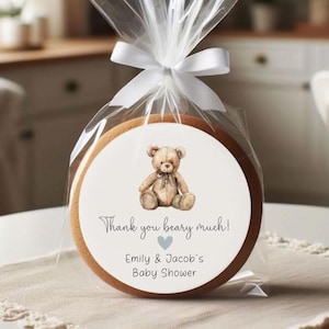 May include: A round, brown cookie with a white sticker featuring a cartoon teddy bear. The sticker says "Thank you beary much! Emily & Jacob's Baby Shower".