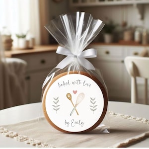 Puede incluir: Una galleta redonda blanca con un borde marr&oacute;n, empaquetada en una bolsa de pl&aacute;stico transparente con una cinta blanca. La galleta tiene una pegatina blanca con el texto "baked with love by Emily" y un coraz&oacute;n rosa, junto con un dibujo de dos utensilios de cocina cruzados.