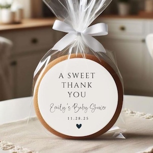 K&ouml;nnte beinhalten: Ein runder wei&szlig;er Keks mit einem schwarz-wei&szlig;en Design, auf dem "A Sweet Thank You Emily's Baby Shower 11.28.25" steht und unten ein kleines schwarzes Herz. Der Keks ist in durchsichtige Cellophanfolie mit einem wei&szlig;en Band verpackt.