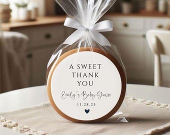 Puede incluir: Una galleta redonda blanca con un diseño en blanco y negro que dice "A Sweet Thank You Emily's Baby Shower 11.28.25" con un pequeño corazón negro en la parte inferior. La galleta está envuelta en celofán transparente con una cinta blanca.