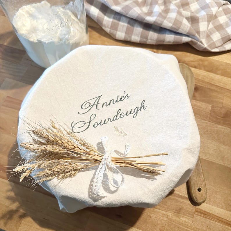 May include: A round, off-white fabric bowl cover featuring the embroidered text "Annie's Sourdough" in a dark grey script font. A small bundle of dried wheat stalks tied with a white lace ribbon is placed on top of the fabric cover.