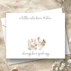 Puede incluir: Una tarjeta blanca con un mensaje escrito a mano que dice "a little note from Katie" y "always be a good egg". La tarjeta presenta una ilustraci&oacute;n de acuarela de dos pollos con flores.
