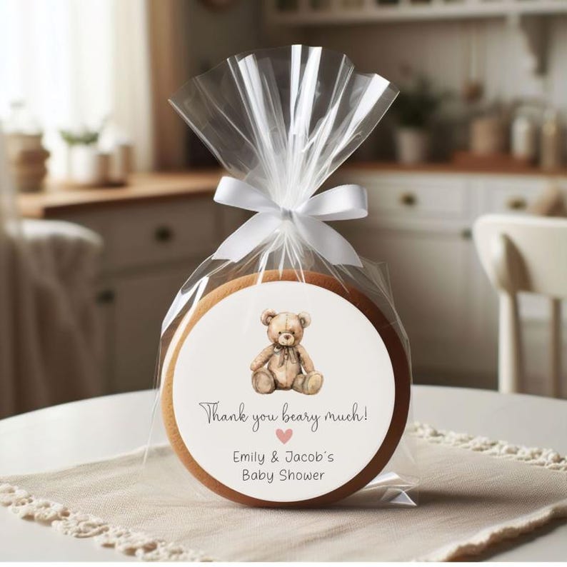 May include: A round, brown cookie with a white sticker featuring a cartoon teddy bear. The sticker says "Thank you beary much!" with a pink heart and "Emily & Jacob's Baby Shower".