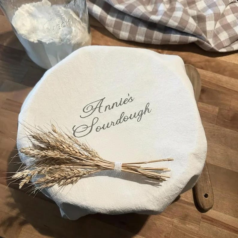 May include: A round, off-white fabric bowl cover featuring the embroidered text "Annie's Sourdough" in a dark script font. A small bundle of dried wheat stalks is tied with white twine and placed across the center of the fabric cover.
