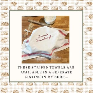 May include: A white kitchen towel with red stripes and the embroidered text Emma's Sourdough, draped over a wooden surface. A wooden rolling pin and a small bundle of wheat stalks are placed nearby, along with a small bowl containing white flour.