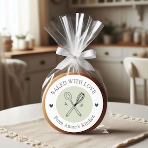 Puede incluir: Una pegatina redonda blanca con el texto "Baked with Love from Annie's Kitchen" y un c&iacute;rculo verde con un batidor y una cuchara en su interior. La pegatina est&aacute; en una galleta marr&oacute;n.