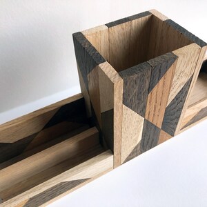 May include: A wooden desk organizer with a geometric pattern in shades of brown and black. The organizer has a tall, square pencil holder and a long, rectangular tray.