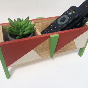 Op de afbeelding: Houten afstandsbediening en plantenhouder. De houder is beschilderd met rode, groene en witte geometrische ontwerpen. Het is verdeeld in twee secties, een voor de plant en de andere voor de afstandsbedieningen.