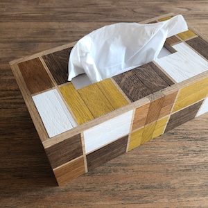 Peut inclure: Un couvre-boîte à mouchoirs en bois avec un motif géométrique de carrés bruns, blancs et jaunes. Le boîte à mouchoirs est ouverte et un mouchoir blanc est partiellement tiré.