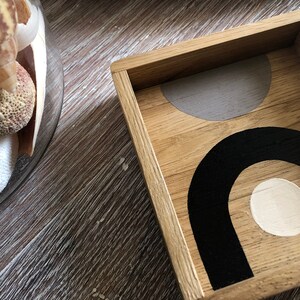 Può includere: Un vassoio di legno con un design geometrico dipinto all'interno. Il design presenta un semicerchio grigio, un arco nero e un cerchio bianco.