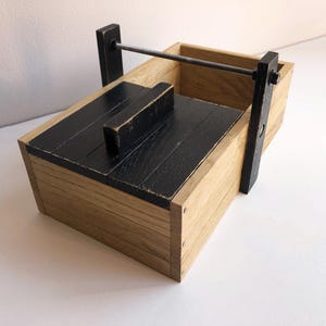Può includere: Una zangola in legno con coperchio e manico neri. La zangola è realizzata in legno chiaro con una finitura scura e usurata sul coperchio e sul manico. Un'asta di metallo si estende sulla parte superiore.