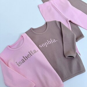 May include: Two long-sleeved shirts, one pink and one brown, with the names "isabella" and "sophia" embroidered on them.