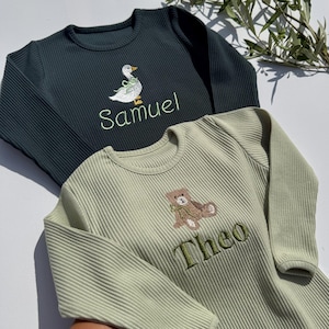 Puede incluir: Dos peleles de bebé de manga larga. Uno es verde oscuro con un bordado de pato blanco y el nombre "Samuel" en verde. El otro es verde claro con un bordado de oso de peluche y el nombre "Theo" en verde. Ambos tienen una textura acanalada.