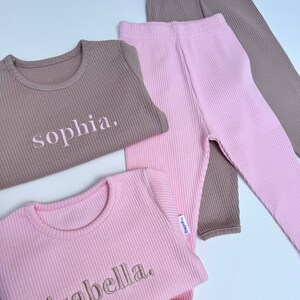 May include: Two sets of ribbed pajamas, one pink and one brown, with the names "sophia" and "isabella" embroidered on the shirts.