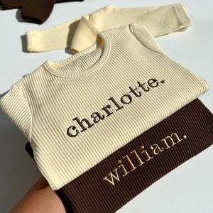 May include: Two ribbed long-sleeve shirts, one cream and one brown, with the names "charlotte" and "william" embroidered in gold thread.