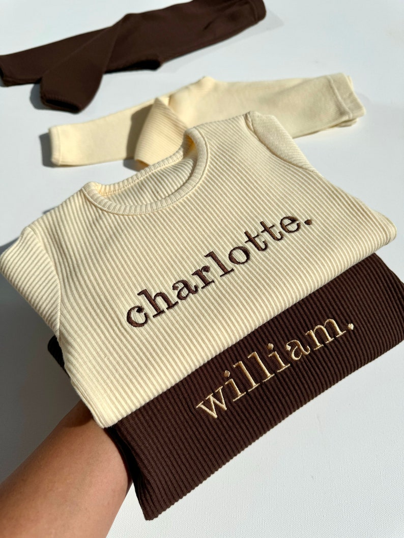 May include: Two ribbed long-sleeve shirts, one cream and one brown, with the names "charlotte" and "william" embroidered in gold thread.