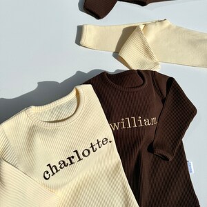 May include: Two long-sleeved shirts, one cream and one brown, with ribbed fabric. The cream shirt has the word "charlotte." embroidered on it, and the brown shirt has the word "william." embroidered on it.