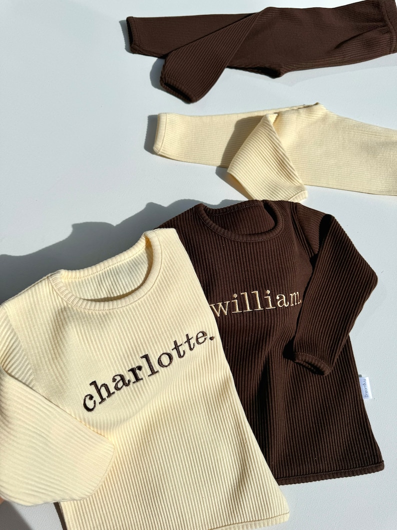 May include: Two long-sleeved shirts, one cream and one brown, with ribbed fabric. The cream shirt has the word "charlotte." embroidered on it, and the brown shirt has the word "william." embroidered on it.
