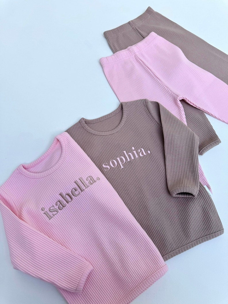 May include: Two long-sleeved shirts, one pink and one brown, with the names "isabella" and "sophia" embroidered on them.