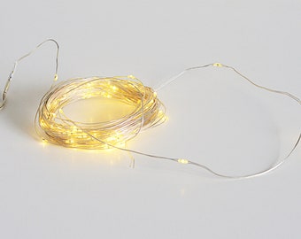 May include: A coiled string of warm white LED fairy lights on a silver wire. The lights are clustered together in a spiral, with some strands extending outward. The lights are on, emitting a soft glow, and are suitable for decorative purposes.