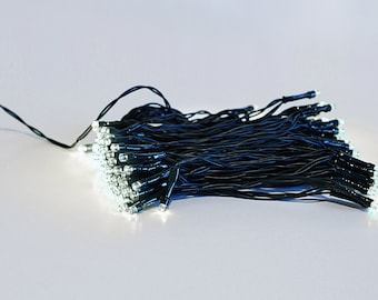 May include: A string of white LED lights on a black wire, illuminated against a white background. The lights are clustered together, with the black wire extending out of the frame. The lights are small and bright.