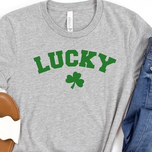 May include: A gray t-shirt with the word "LUCKY" in green letters and a green shamrock underneath. The shirt is paired with blue jeans and white sandals.
