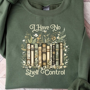 May include: A dark green sweatshirt with the text "I Have No Shelf Control" in a decorative font. The design features a stack of books surrounded by floral elements. The sweatshirt has long sleeves and a crew neck.