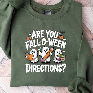 May include: A green sweatshirt with the text "ARE YOU FALL-O-WEEN DIRECTIONS?" and illustrations of ghosts, a pencil, a book, a pumpkin, and a bat. The sweatshirt is folded on a light wood-grain surface.