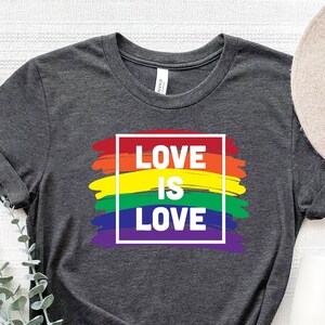 May include: Dark gray t-shirt with the text "LOVE IS LOVE" in a white outlined box, set against a rainbow-colored brushstroke design. A neutral-toned hat and white mug are next to the shirt.
