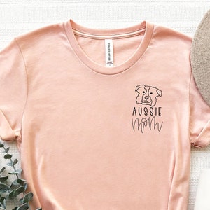 May include: A peach-colored t-shirt with a graphic of an Australian Shepherd dog and the text "Aussie Mom" on the front.