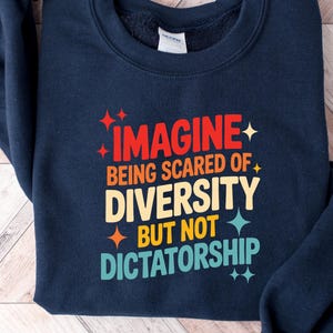 May include: Navy blue sweatshirt with the text "IMAGINE being scared of DIVERSITY BUT NOT DICTATORSHIP" in red, yellow, and teal. The text is surrounded by starburst graphics. The sweatshirt is folded on a light wood grain surface.